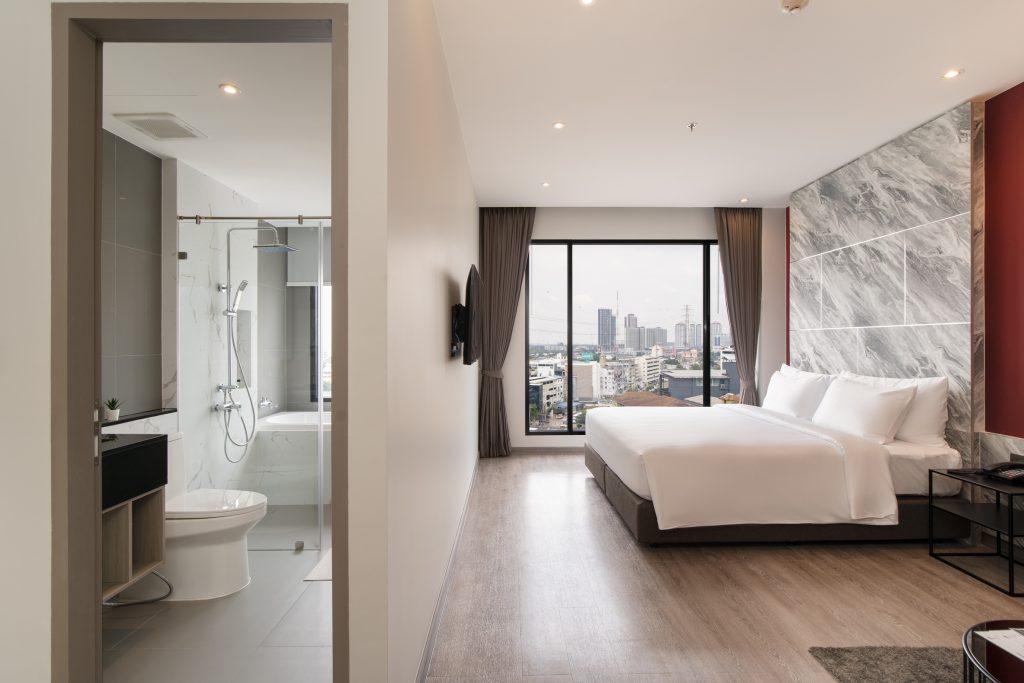 Modern hotel room with a large bed, marble accent wall, floor-to-ceiling window with city view, and an open bathroom featuring a glass shower and toilet. The space is bright and neatly arranged.
