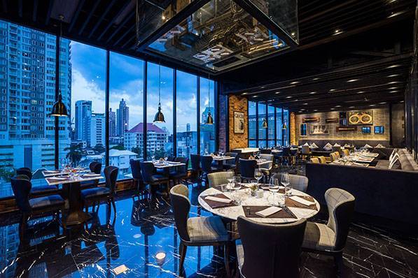 Modern upscale restaurant with elegant tables and chairs, large windows offering a city skyline view at dusk, dark polished floors, ambient lighting, and stylish decor.