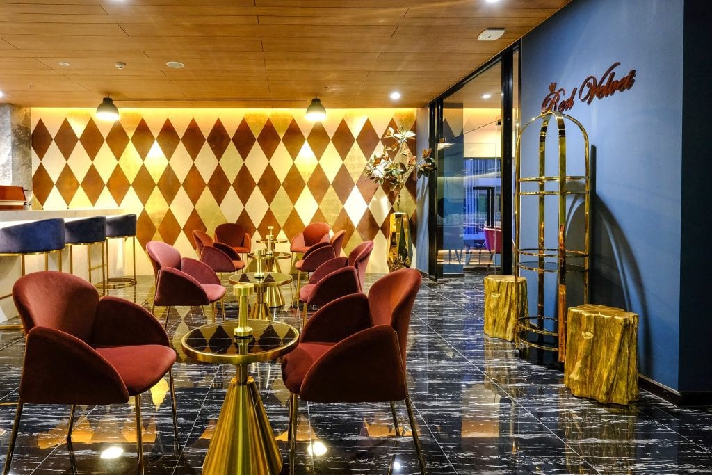 A stylish lounge with maroon chairs, gold tables, a geometric gold and white accent wall, black marble floors, and a blue wall with Red Velvet signage. A gold luggage cart and decorative plants are also visible.