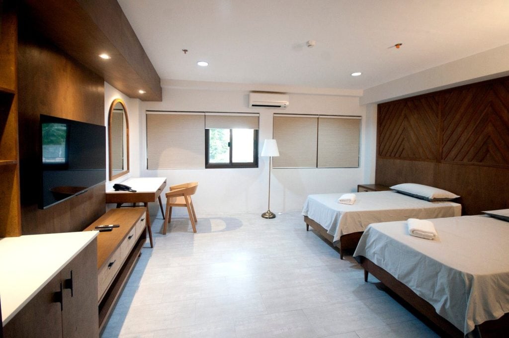 A modern hotel room with two single beds, white bedding, towels, a wooden desk and chair, a flat-screen TV, a standing lamp, and large windows with blinds. The decor features warm wood tones.