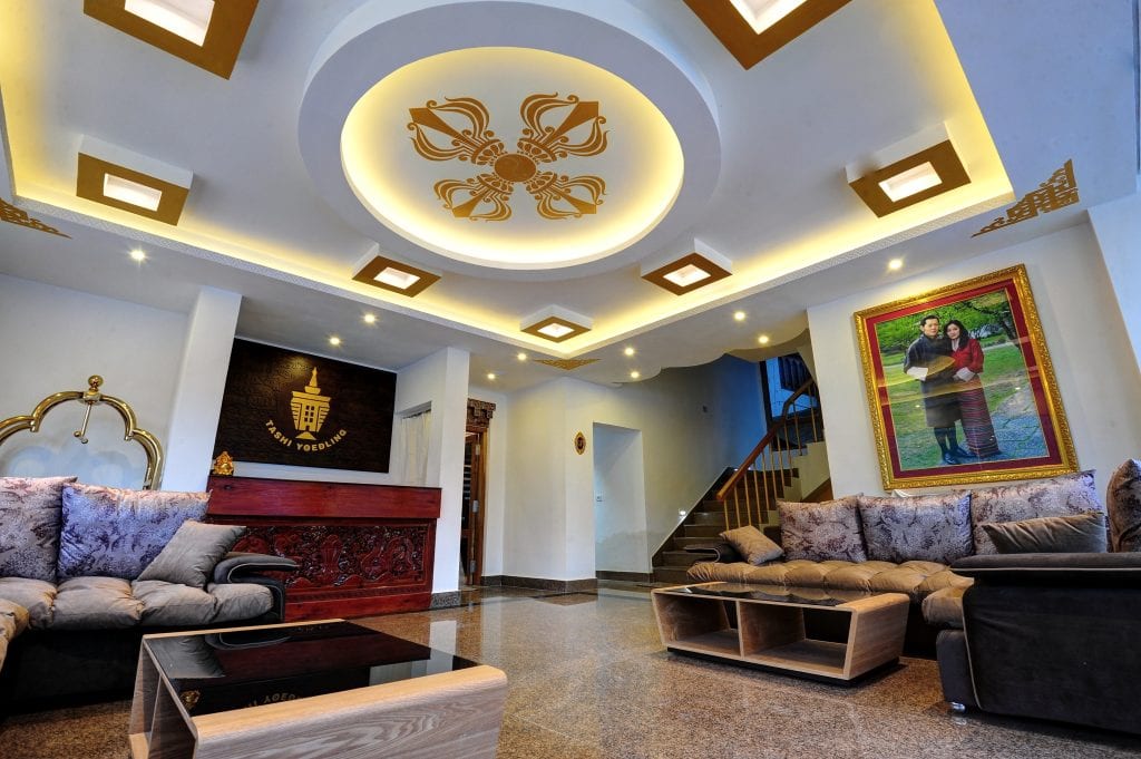 A modern living room with plush sofas, a glossy floor, a black and wood coffee table, and a red wooden counter. The ceiling has ornate gold decorations and bright recessed lighting. A large framed portrait hangs on the wall.