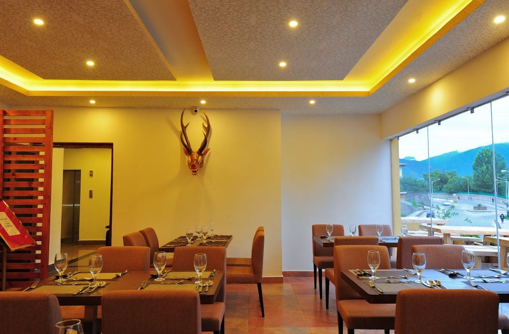 A modern restaurant dining area with neatly set tables and chairs, a decorative antler mount on the wall, large windows offering a view of mountains and a road outside, and warm ceiling lights illuminating the space.