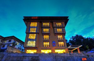 A brightly lit four-story building with traditional Bhutanese architectural details stands against a deep blue evening sky, with illuminated windows and decorative patterns around the edges.