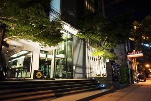 Nighttime street view of a modern building with large glass windows and bright lighting, surrounded by trees. A lit sign on the right reads MARIGOLD. The sidewalk and street are quiet and empty.