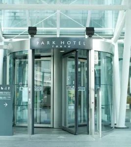 A glass revolving door entrance to Park Hotel Tokyo, with the hotel name displayed above and a sign with information on the left side. The modern building features transparent glass and metallic frames.