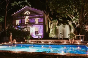 A brightly lit old stone house stands by a glowing pool at night, surrounded by lush trees. A waterfall cascades beside the house, with soft lights illuminating the scene and potted plants along the pool’s edge.