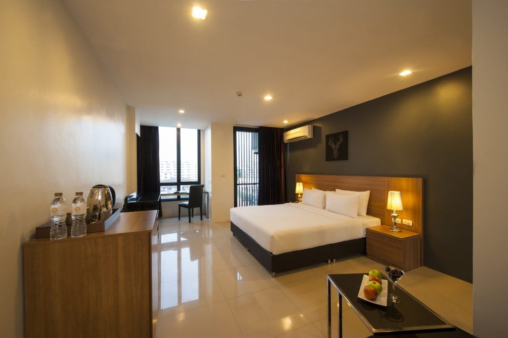 Modern hotel room with a large bed, two bedside tables with lamps, a desk by the window, and a dresser with bottled water and a kettle. A small table with fruit and drinks sits in the foreground.