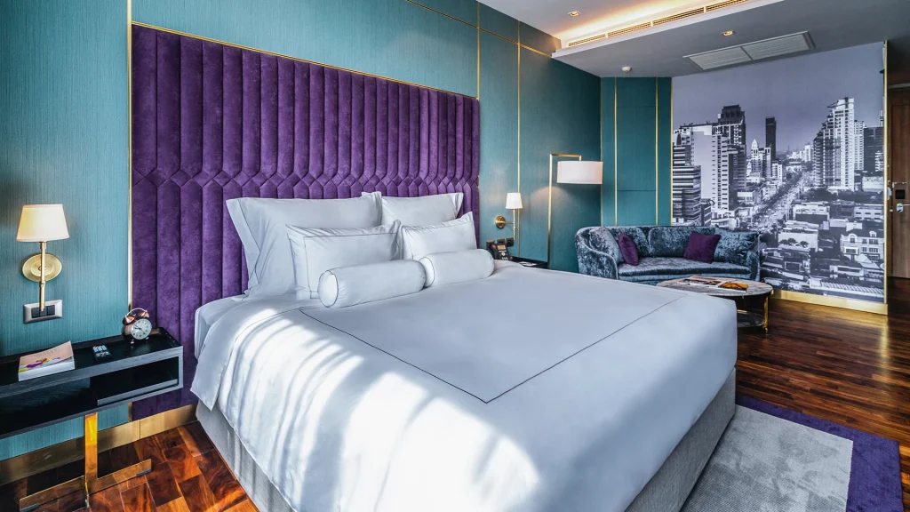 Modern hotel room with a large bed, white bedding, and a tall, purple tufted headboard. The walls are teal, and there’s a cityscape mural, a gray sofa, lamps, a side table, and wood flooring.