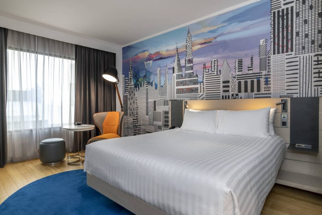 Modern hotel room with a large bed, white striped bedding, and a cityscape mural behind the headboard. The room features a window with sheer and dark curtains, a yellow chair, floor lamp, small round table, and hardwood floors.