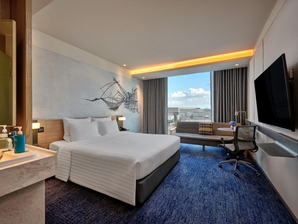 Modern hotel room with a king-sized bed, wall art of a ship, blue carpet, large window with city and sea view, a desk with a chair, a sofa, and a mounted TV; toiletries on a counter in the foreground.