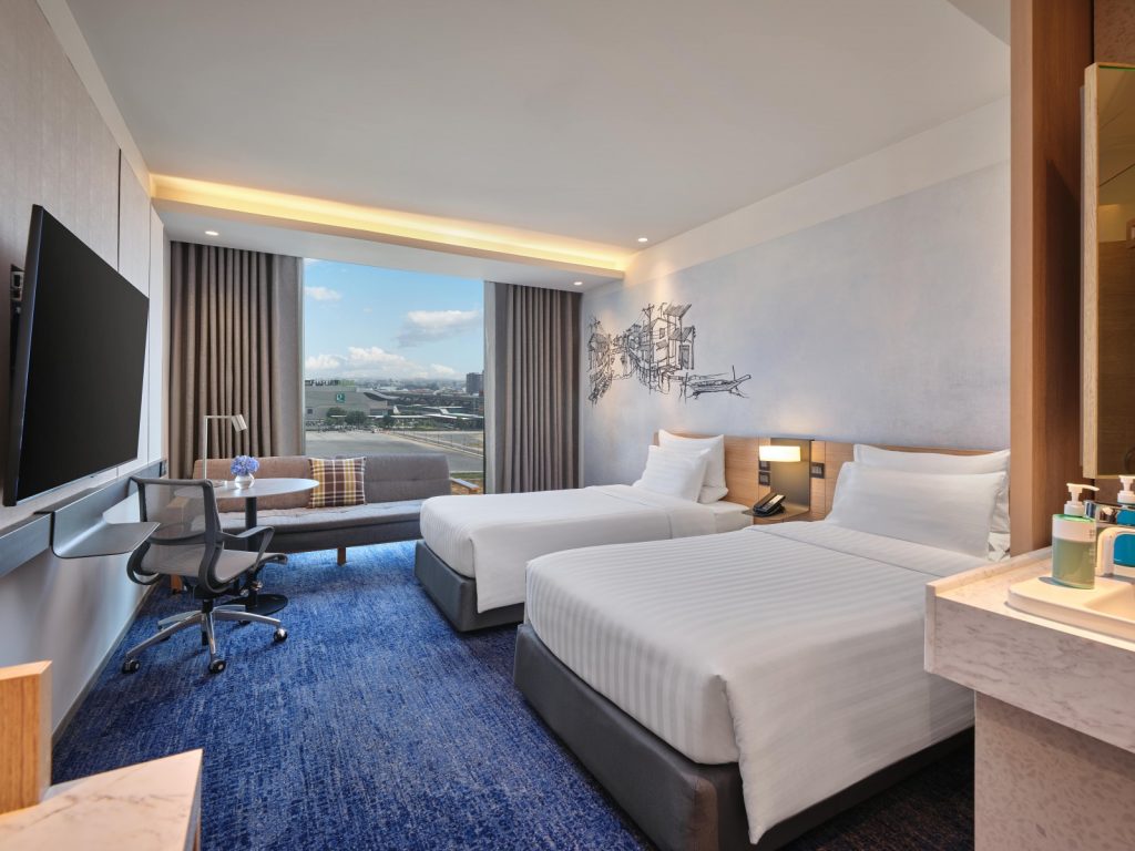Modern hotel room with two twin beds, a large flat-screen TV, a work desk with a chair, a lounge area by a window with city views, and a clean, contemporary design featuring blue and neutral tones.