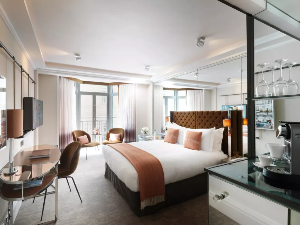 Modern hotel room with a large bed, brown and white bedding, two brown chairs by a window, a desk with a chair, and a glass shelf with glasses and a coffee machine. Light fills the room through large windows.