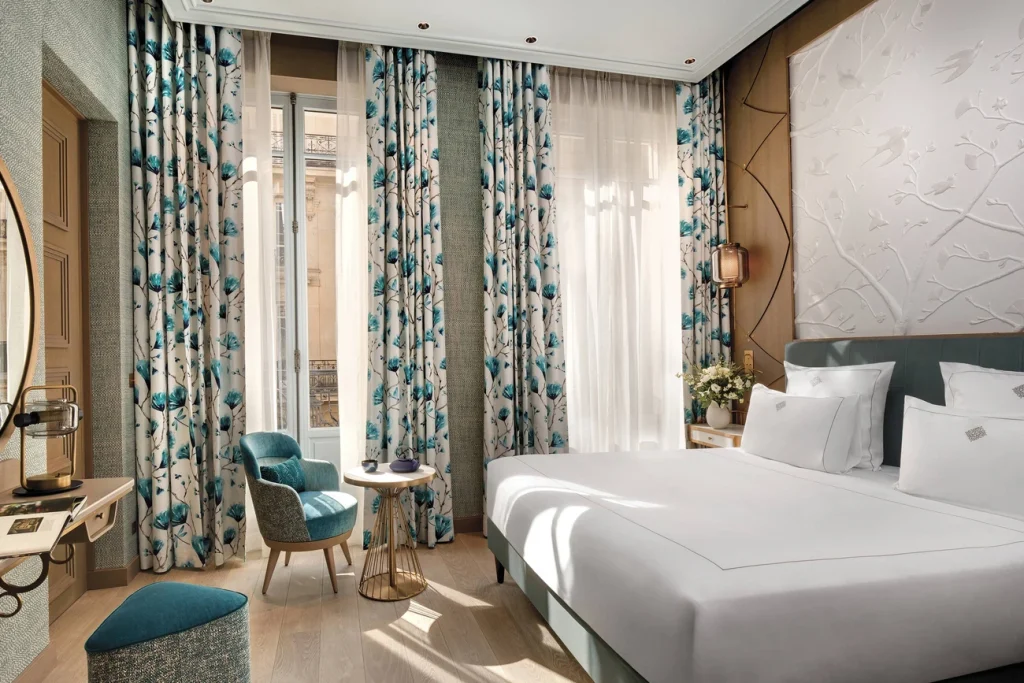 Elegant hotel room with a large bed, blue and white floral curtains, a small table with chairs, soft lighting, and a wall panel with a nature design. Sunlight streams in through tall windows.
