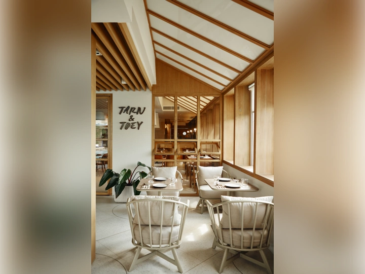 A cozy, modern café with wooden beams, large windows, and natural light. Cream cushioned chairs surround small tables, and a plant sits near a wall with the sign TARN & TOEY. The atmosphere is warm and inviting.