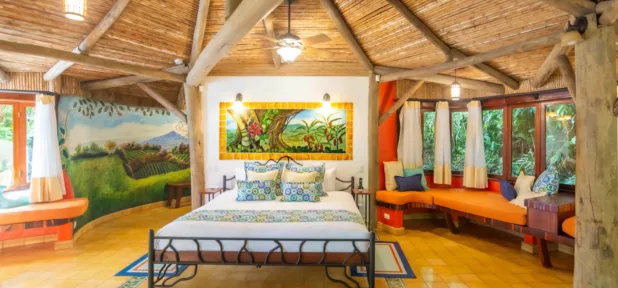 A cozy bedroom with a rustic wooden ceiling, colorful artwork above the bed, large windows with curtains, built-in window seats with cushions, and warm yellow-tiled floors, surrounded by lush greenery outside.