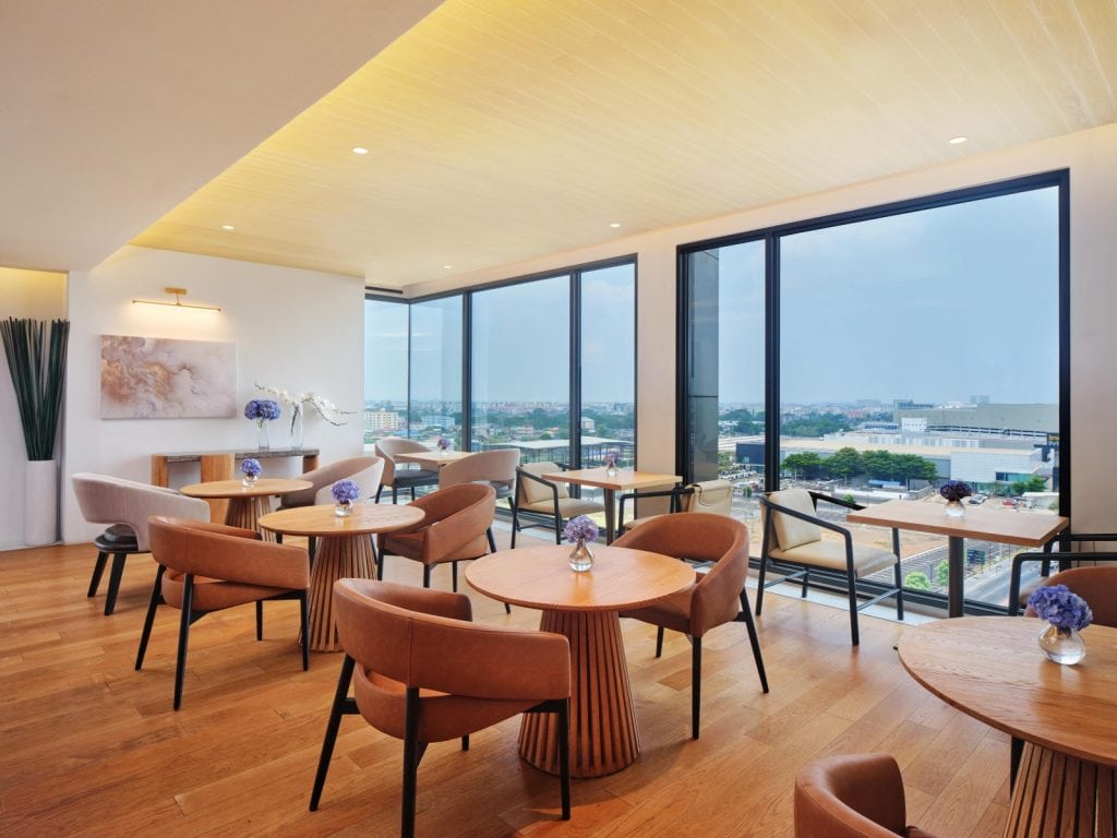 A modern, elegant café with wooden floors, round wooden tables, brown chairs, large windows offering a city view, and small vases with purple flowers on each table. Natural light fills the bright, spacious room.