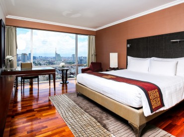 A modern hotel room with a large bed, white linens, a desk, a chair, and floor-to-ceiling windows offering a city skyline view. The room has wooden floors and contemporary decor.