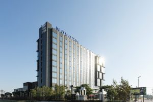 A tall, modern Novotel hotel building with many windows, reflecting sunlight. The hotel sign is visible at the top, and there are trees and a small statue in front, under a clear blue sky.