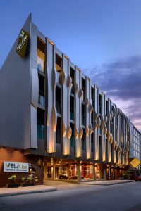 Modern hotel building at dusk with unique, wavy vertical design elements on the facade. Warm lights illuminate the exterior, and signs read VELA be above the entrance and on the corner of the building.