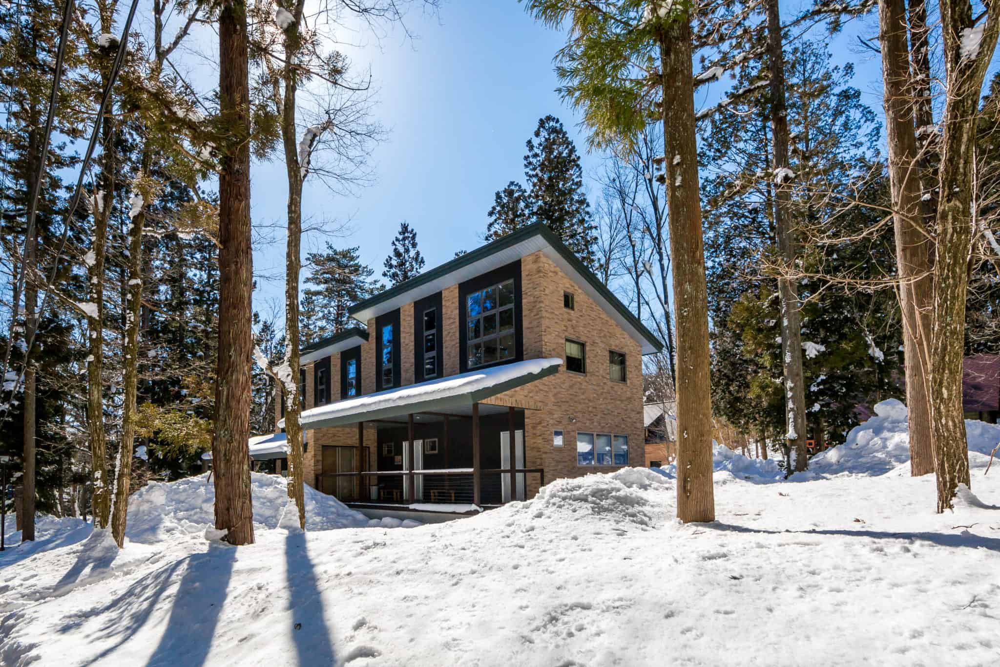 A modern two-story house with large windows and a slanted roof stands amid tall trees and deep snow under a clear blue sky. The sunlight casts shadows on the snow-covered ground surrounding the home.