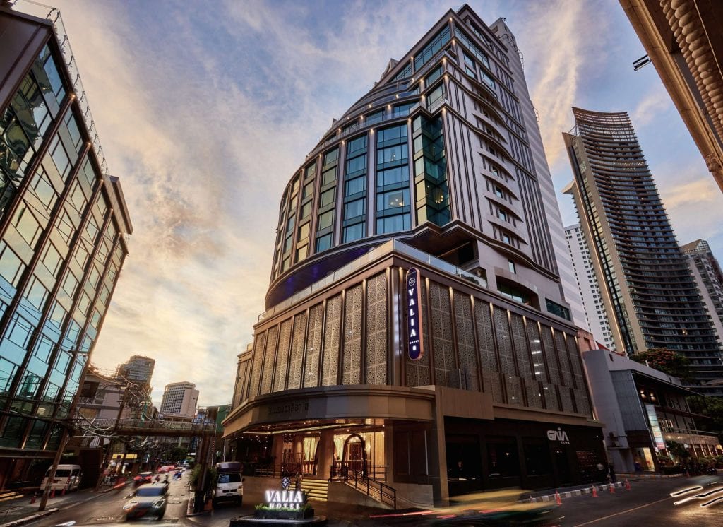A modern high-rise hotel with large glass windows and a sign reading VALIA is set against a sunset sky, surrounded by other tall buildings and light traffic on the street below.