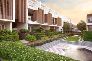 Modern townhouse buildings with large windows and wooden panel accents overlook landscaped gardens, manicured hedges, and a patterned courtyard at sunset.