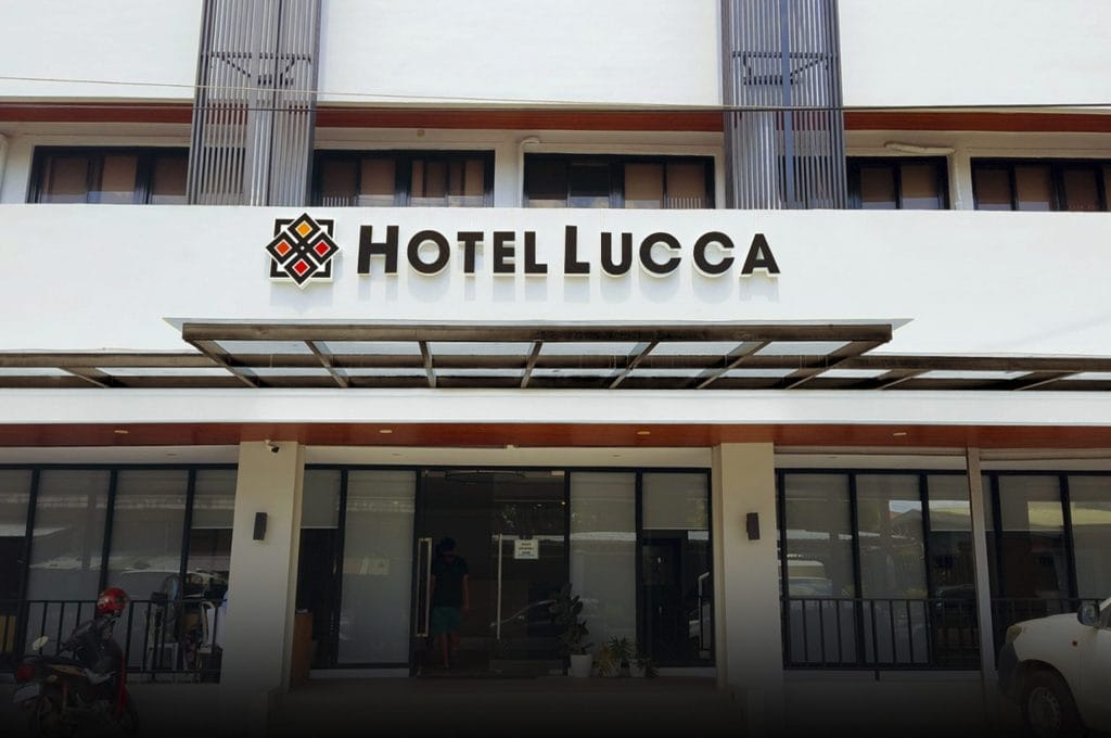 The front entrance of Hotel Lucca, featuring a modern white exterior, a glass canopy, and a large black, white, and red hotel sign above the doorway. A person stands at the reception area inside.