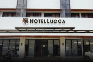 The front entrance of Hotel Lucca, featuring a modern white exterior, a glass canopy, and a large black, white, and red hotel sign above the doorway. A person stands at the reception area inside.