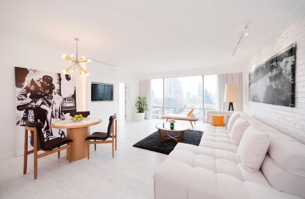 A modern, bright living room with white walls and furniture, a large sectional sofa, round dining table, contemporary decor, and large windows offering a city skyline view.