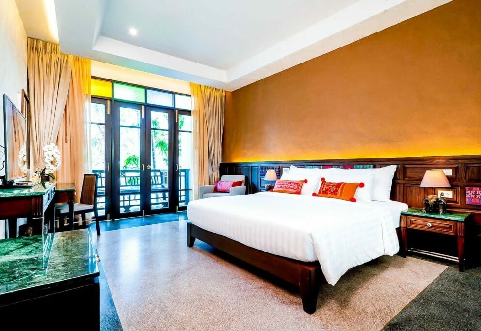 A bright hotel room with a large bed, white linens, colorful pillows, a sofa, and a desk. Sunlight streams through glass doors leading to a balcony with greenery outside. The walls are tan and dark wood.