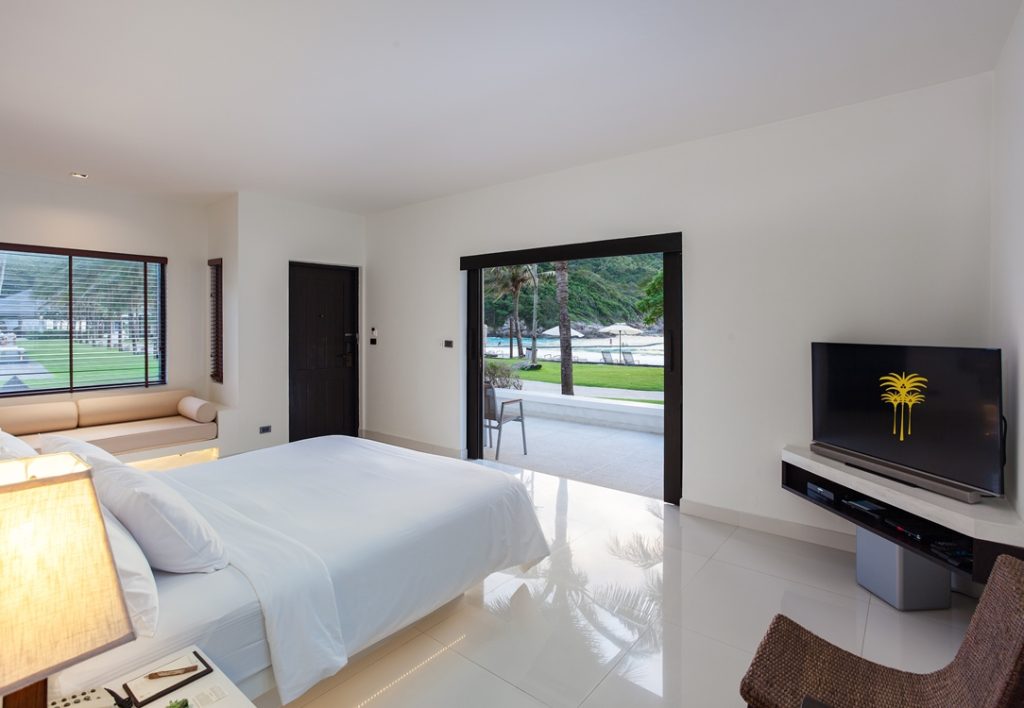 Modern hotel room with a large bed, flat-screen TV, and sliding glass doors opening to a patio with outdoor seating and a view of greenery and hills. Natural light fills the bright, minimalist space.