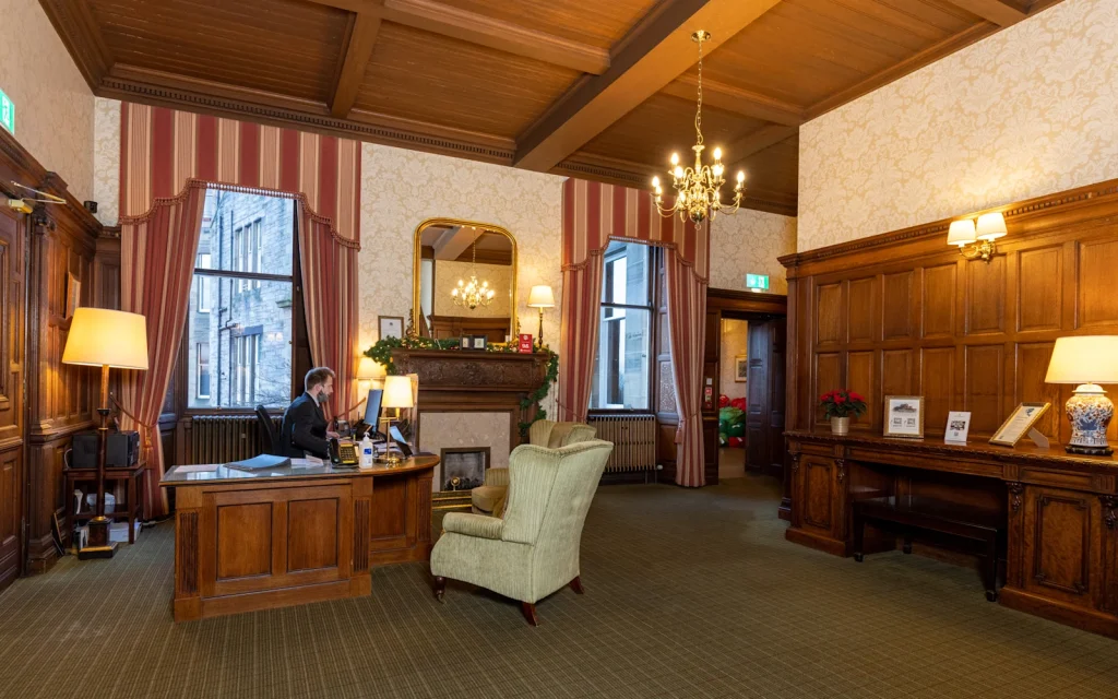 A receptionist sits at a wooden desk in an elegant, traditional hotel lobby with wood paneling, striped wallpaper, a fireplace, chandeliers, large windows, and a light green armchair.