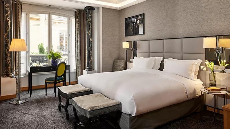Elegant hotel room with a large bed, two benches at the foot, a desk with a chair by the window, lamps, and modern decor. Light streams through tall French windows with curtains, and fresh flowers are on the desk.