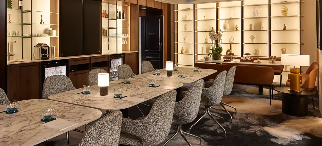 A modern lounge with marble tables, gray chairs, soft lighting, and glass shelves displaying art pieces and vases. There are table lamps, decorative items, and a cozy, elegant ambiance.
