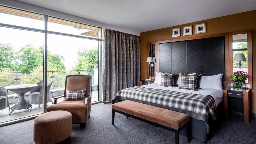A modern hotel room with a large bed, plaid pillows and throws, a padded bench, and a sitting area. Glass doors open to a balcony with outdoor furniture and views of trees. The decor is neutral with warm accents.