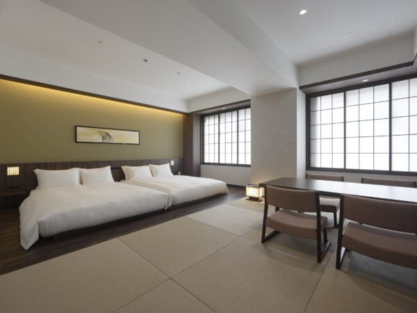 A modern Japanese-style hotel room with two beds, a low dining table with two chairs, tatami mat flooring, shoji screen windows, and minimalistic decor. Soft lighting creates a calm and inviting atmosphere.