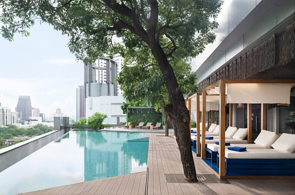 A modern rooftop infinity pool with city views, wooden deck, shaded cabana beds with blue cushions, and a large tree providing shade. Tall buildings are visible in the background.