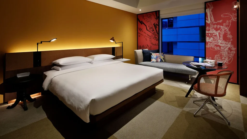 Modern hotel room with a large bed, two reading lights, a gray sofa by the window, a desk with a chair, and contemporary wall art. The window shows a blue-lit building outside at dusk.