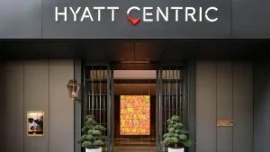 Hyatt Centric hotel entrance with modern gray facade, illuminated sign above doors, potted plants on either side, and a warm, colorful geometric art piece visible inside the lobby.