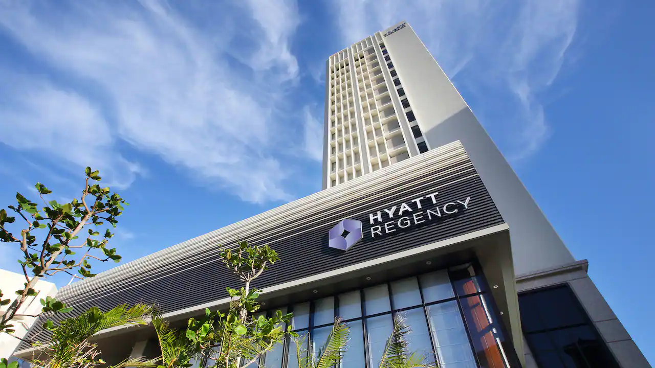 A tall Hyatt Regency hotel building viewed from below, with a modern facade, large windows, and the hotel logo visible. Trees and blue sky with wispy clouds are in the background.