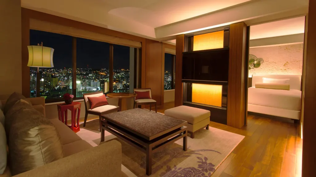 A modern hotel suite at night with a cozy living area, contemporary furniture, warm lighting, and large windows offering a city skyline view. A partition separates the bedroom, where a bed is visible.