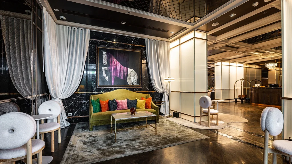 A modern, elegant hotel lobby with a green velvet sofa, colorful pillows, white round chairs, a marble floor, sheer curtains, and a large abstract painting on a black marble wall. A reception desk is visible in the background.