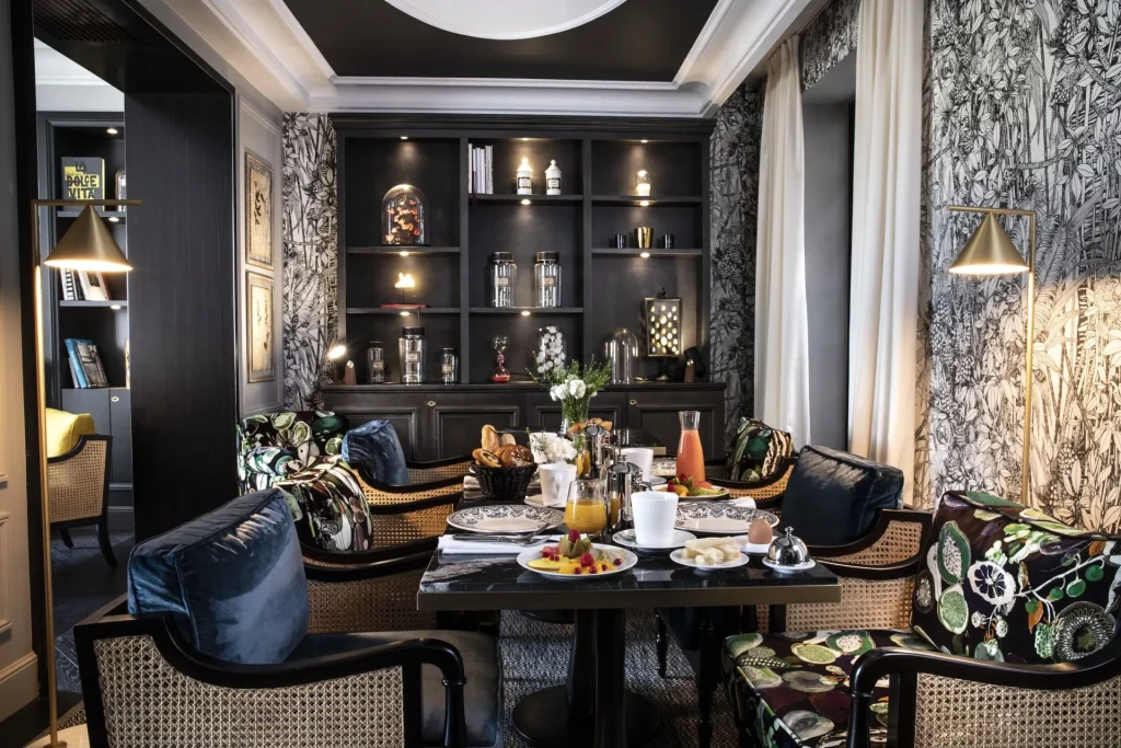 A stylish dining room with a black bookshelf, patterned wallpaper, and a set table with breakfast foods, drinks, and pastries. Four cushioned chairs with floral prints surround the table. Warm lighting creates a cozy atmosphere.