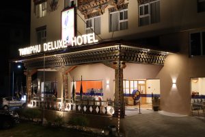 Night view of the Thimphu Deluxe Hotel entrance, brightly lit with ornate wooden carvings, flags along the front, and parked cars visible to the left.