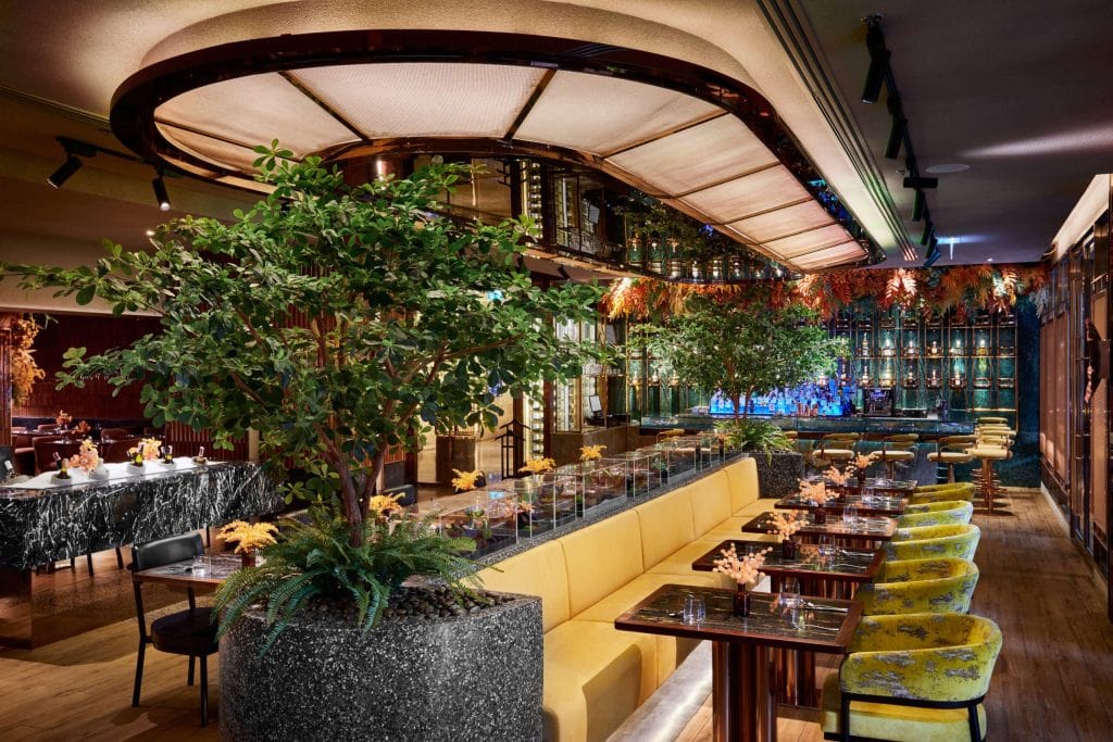 A stylish restaurant interior with modern decor, large potted plants, yellow cushioned seating, marble tables, velvet chairs, and a decorative wall with shelves and greenery, all warmly lit for an inviting atmosphere.