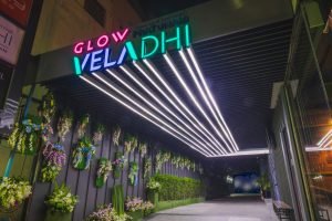 An entrance hallway with glowing neon signs reading GLOW VELADHI and linear ceiling lights. The walls are decorated with green plants and flowers arranged vertically.