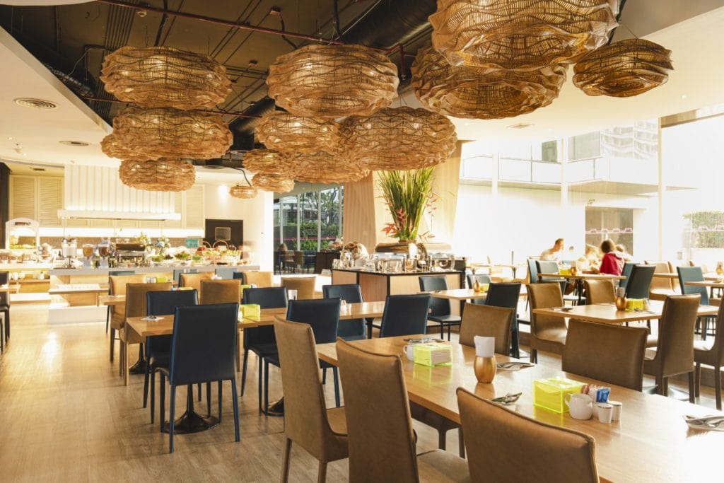Modern restaurant interior with wooden tables, brown and navy chairs, large woven pendant lights, and floor-to-ceiling windows letting in natural light. A buffet area is visible in the background, with a few people dining.