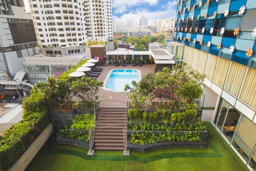 View of a modern hotel courtyard with a rectangular swimming pool, poolside lounge chairs, a rooftop garden with lush greenery, and tall city buildings in the background under a partly cloudy sky.
