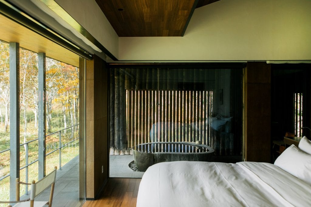 A modern bedroom with white bedding features a large window overlooking trees and a balcony. A free-standing bathtub sits in front of floor-to-ceiling glass, and natural light fills the room.