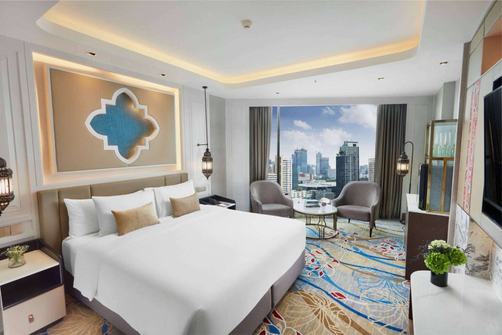 A modern hotel room with a large bed, bedside tables, two chairs and a round table by a window overlooking a city skyline. The room features decorative lighting, patterned carpet, and contemporary decor.